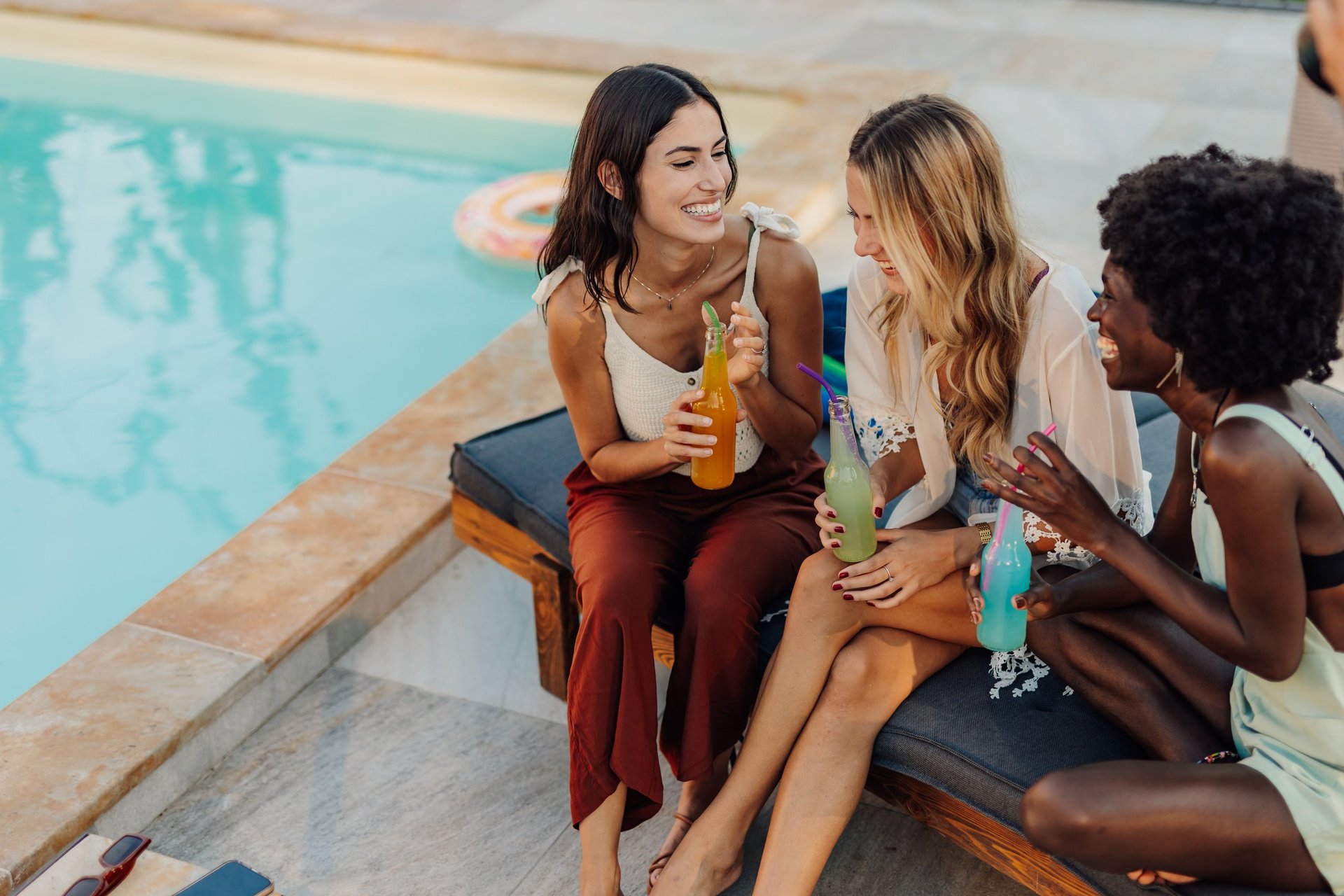 Three multi-ethnic female friends are enjoying their vacation, sitting by the pool and drinking cocktails