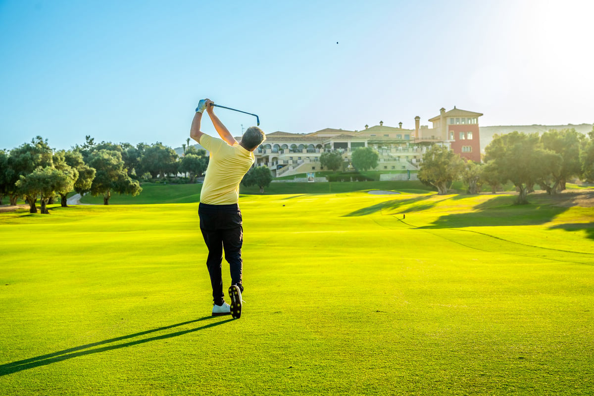 Mature male golfer swinging his club on a lush green golf course, with a luxurious clubhouse in the background