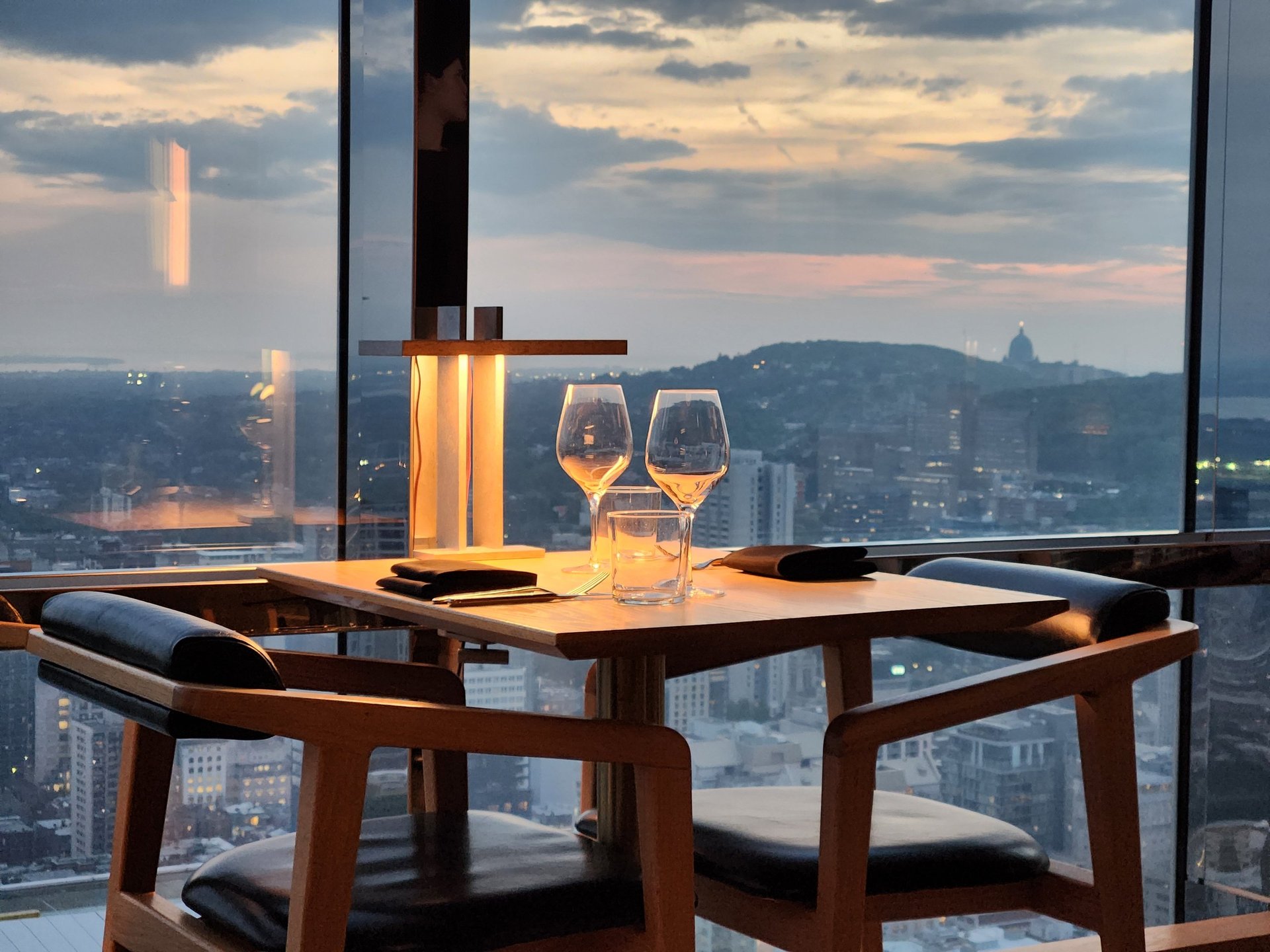 Elegant restaurant table set with wine glasses and soft ambient lighting near a large window overlooking a city skyline at sunset