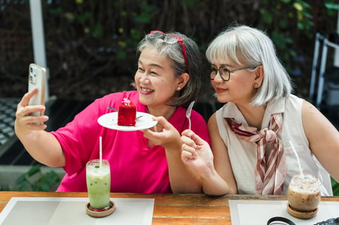 Senior Asian women hold cake and smartphone for selfie at cafe, smiling during sweet reunion moment