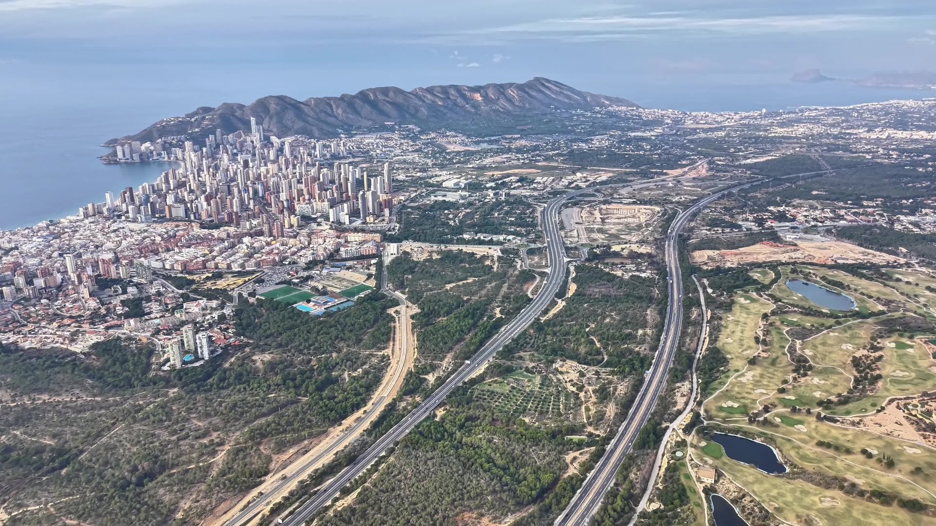 Golf courses and urban skyline from above with winding fairways, green lakes, highways and distant ocean view