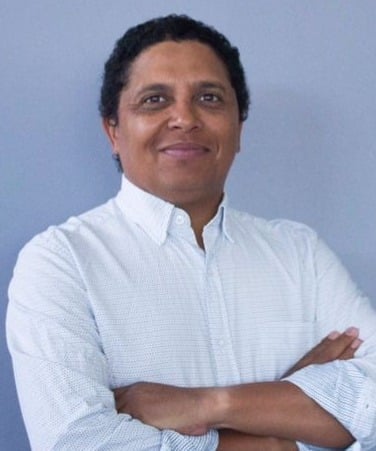 Professional portrait of a man wearing a white button-up shirt, arms crossed, smiling against a light blue background.